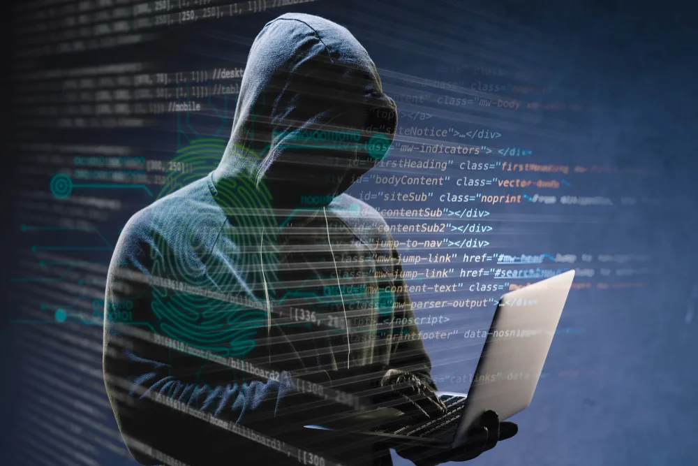 Hooded figure with a laptop behind lines of code